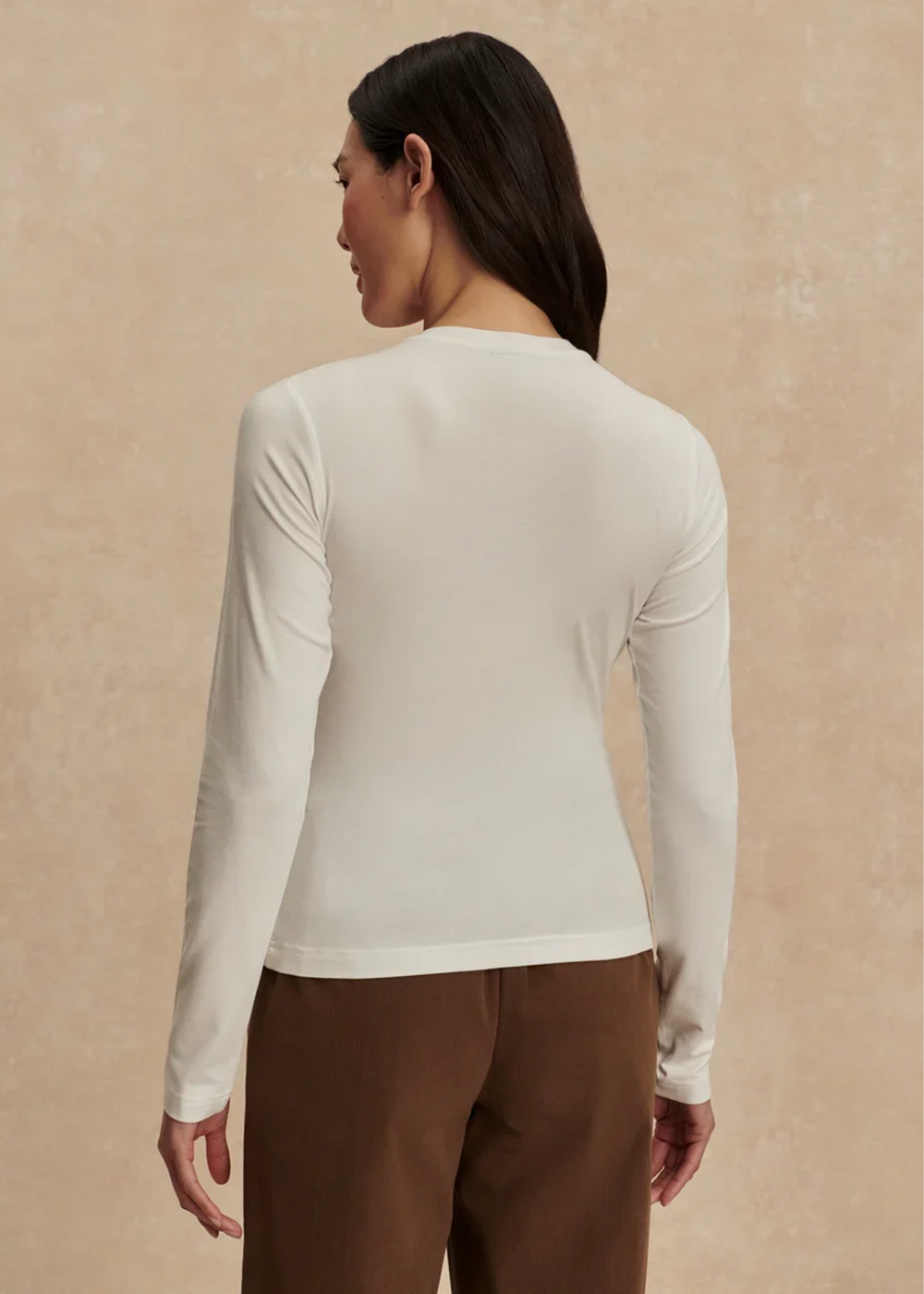 A woman shown from the back wearing a fitted long-sleeve crew-neck top in off-white and brown pants, standing against a warm beige textured studio background.