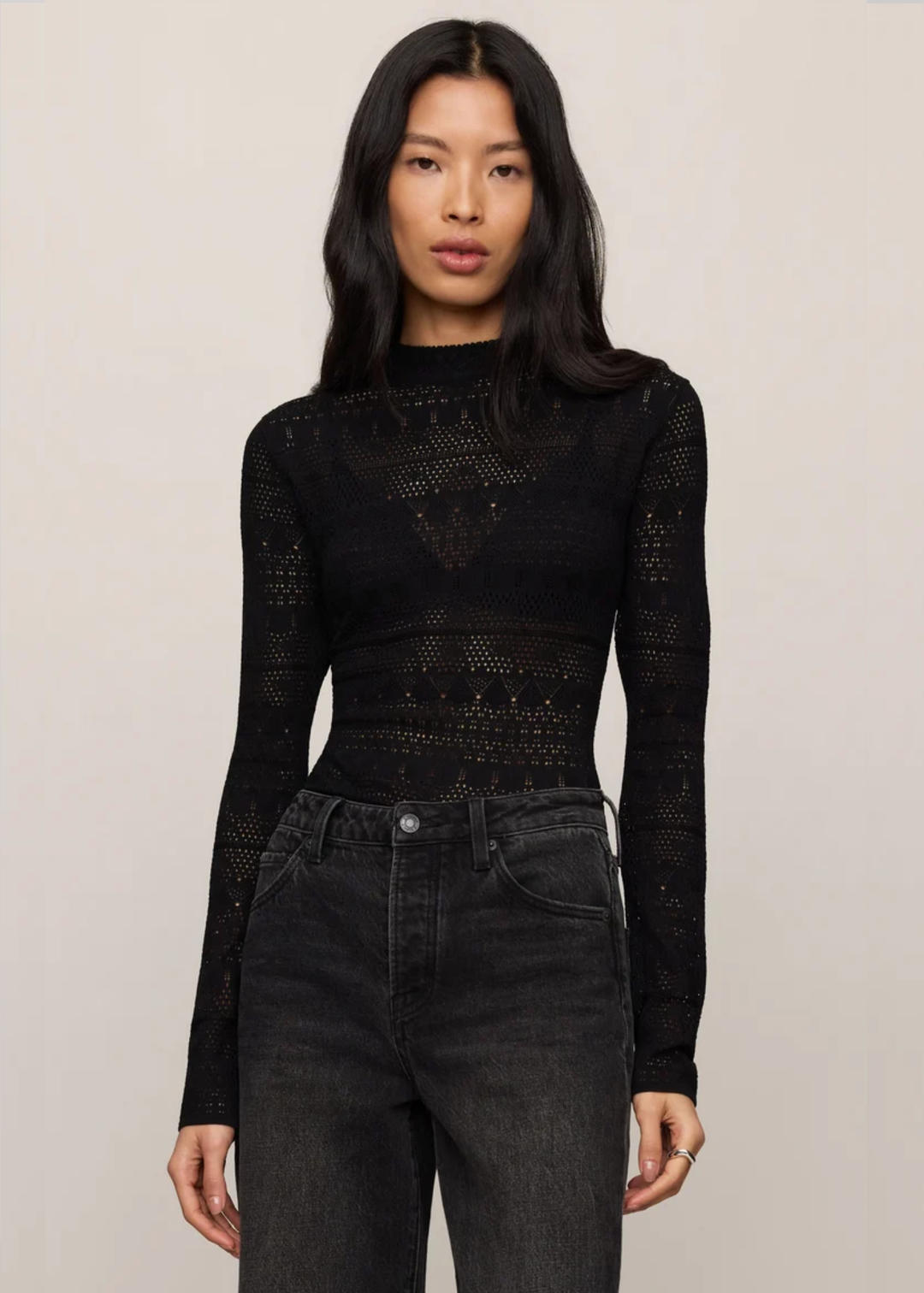 A woman modeling a black long-sleeve knit top with a textured, openwork geometric pattern, paired with dark gray jeans, against a plain light beige background.