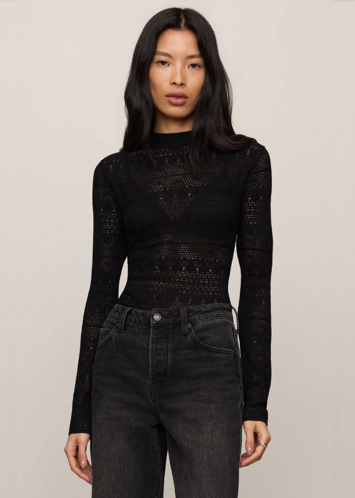 A woman modeling a black long-sleeve knit top with a textured, openwork geometric pattern, paired with dark gray jeans, against a plain light beige background.