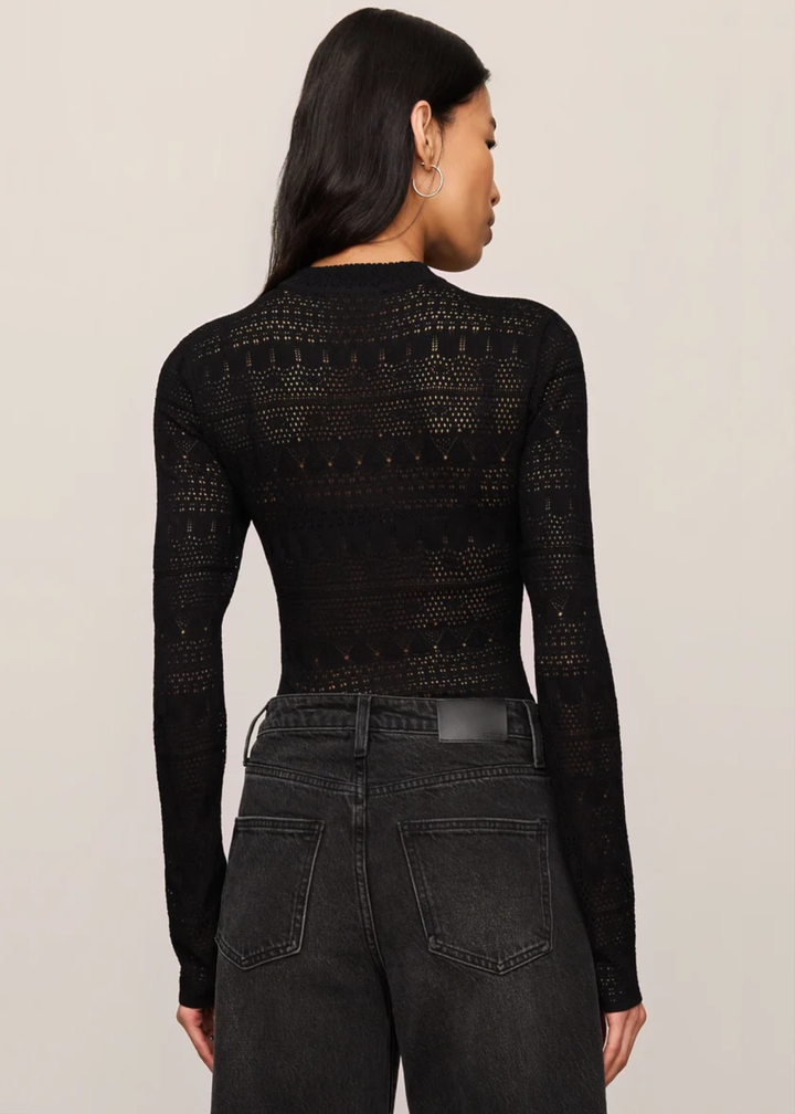 A woman shown from the back wearing a black long-sleeve knit top with a textured openwork pattern, paired with dark gray jeans, against a plain light beige studio background.