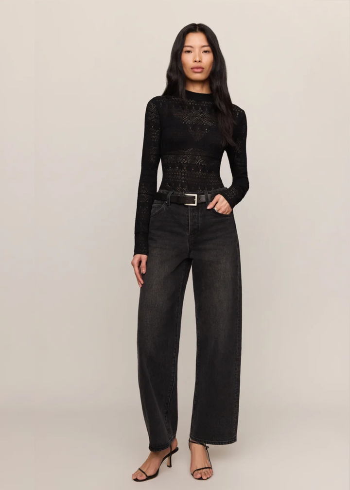 A model stands against a plain light beige studio background wearing a black long-sleeve lace-trim top, black wide-leg jeans with a black belt, and black heeled sandals.