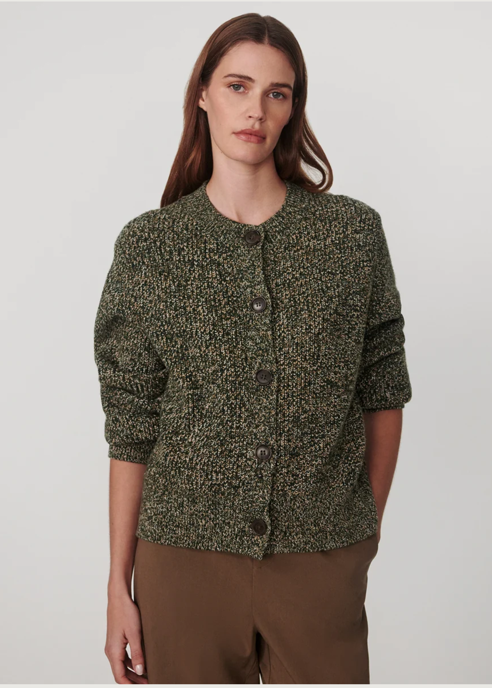 A woman modeling a textured, speckled green and black button-up cardigan sweater with brown pants, shot against a neutral light gray background.