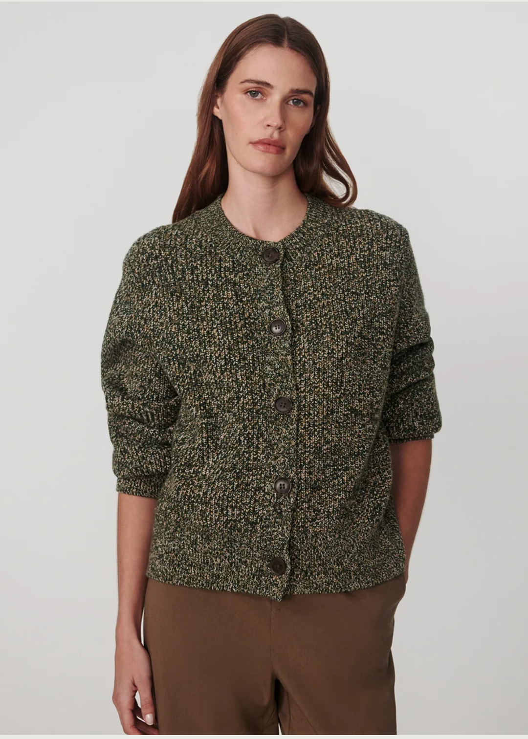 A woman modeling a textured, speckled green and black button-up cardigan sweater with brown pants, shot against a neutral light gray background.