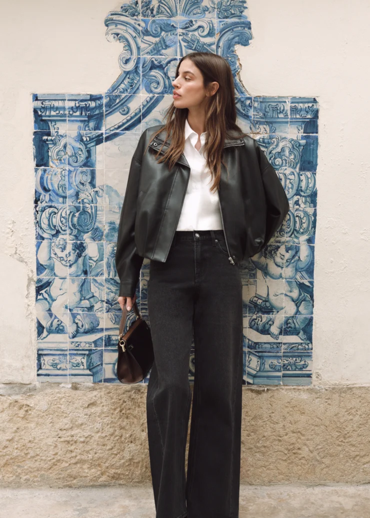 A woman stands outdoors against a white wall with blue ceramic tile panels, wearing a black leather jacket over a white collared shirt and black wide-leg jeans, holding a small dark handbag.