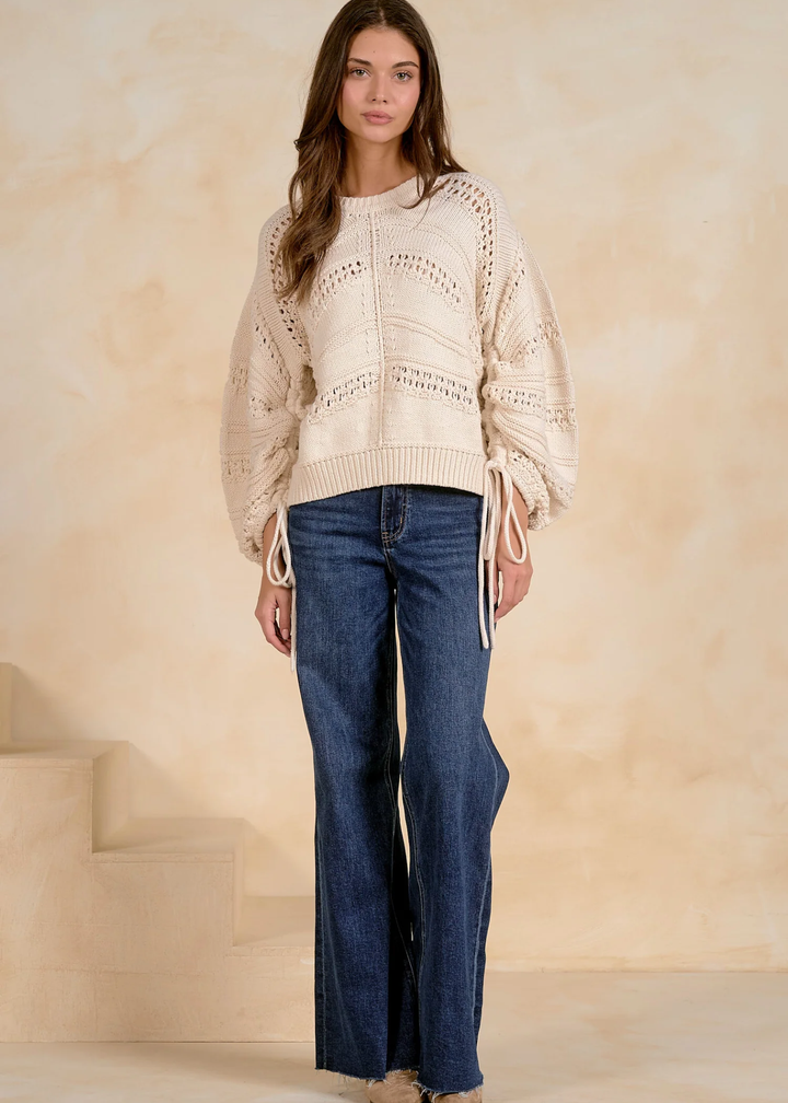 A woman modeling a cream-colored open-knit sweater with balloon sleeves and side tie details, paired with dark blue wide-leg jeans, set against a warm beige textured wall and steps.