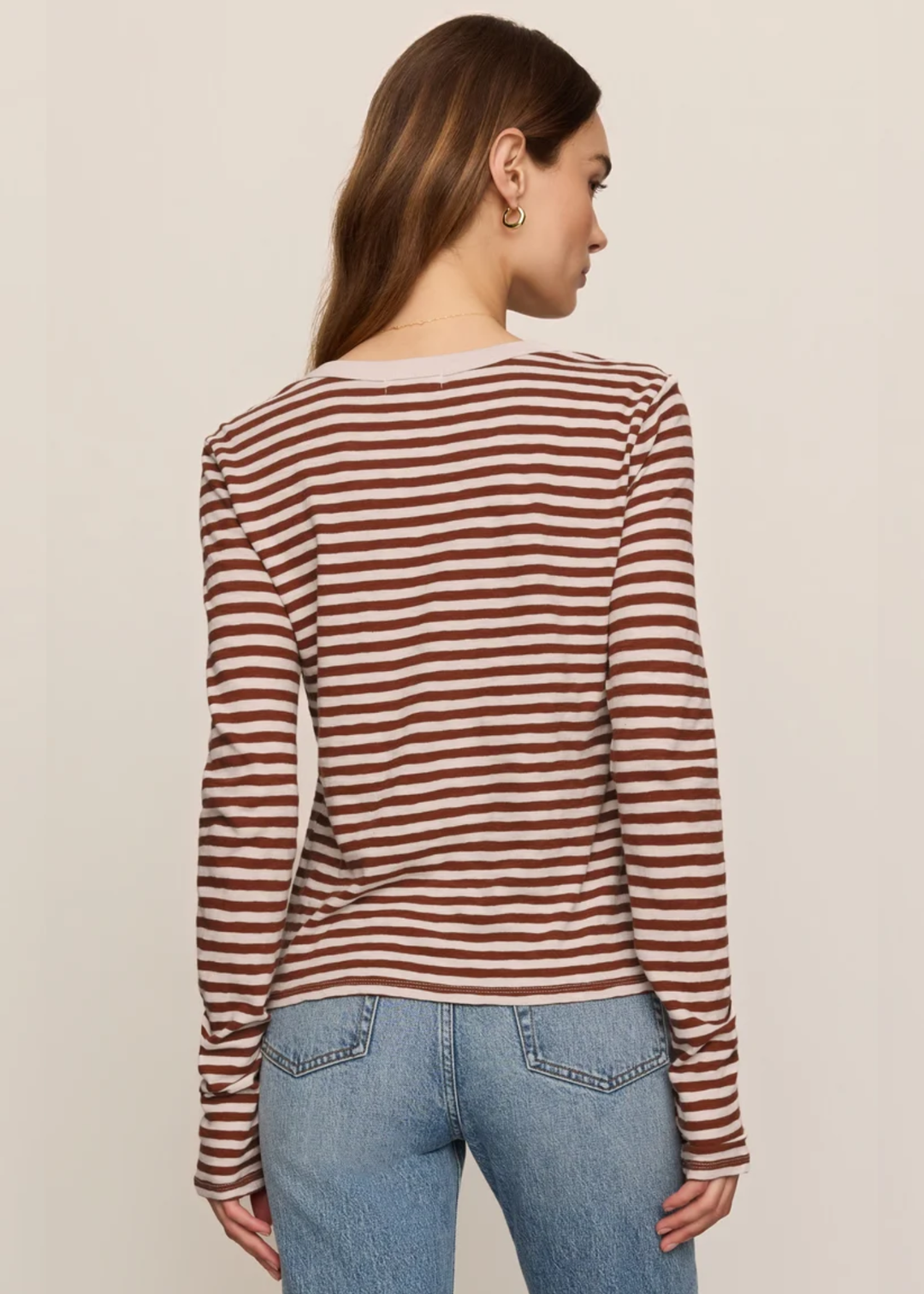 A woman shown from the back wearing a long-sleeve ribbed top with horizontal rust-brown and off-white stripes, paired with light blue jeans, against a plain light beige studio background.