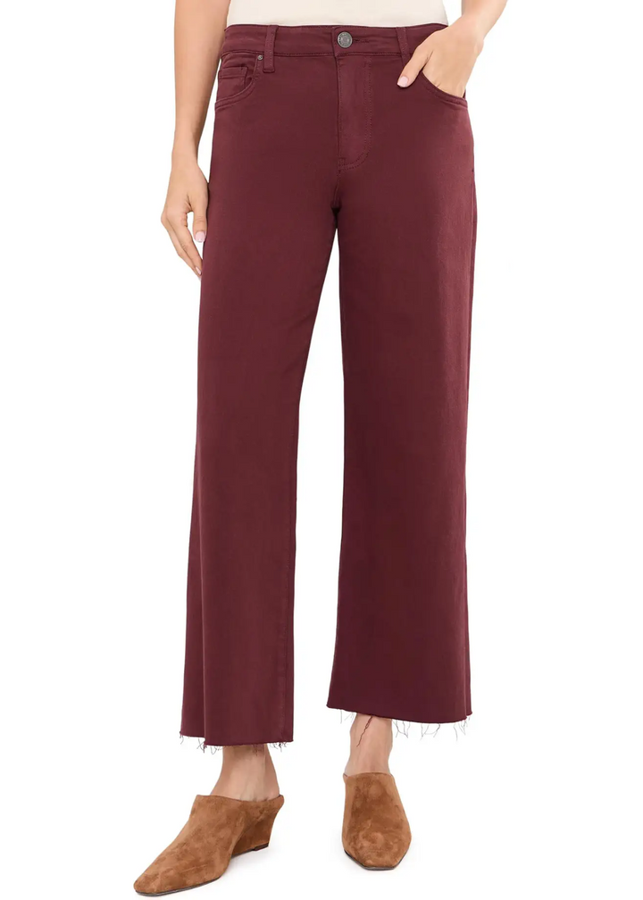 A model wearing a pair of wide-leg cropped jeans in a deep burgundy color, styled with brown suede wedge mules against a plain white background.