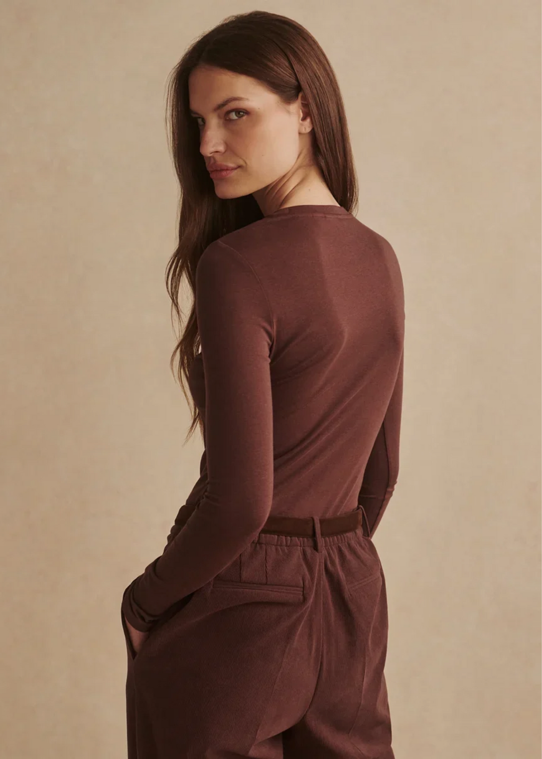 A woman modeling a fitted long-sleeve crewneck top in a deep warm brown, paired with matching brown trousers and a brown belt, against a plain beige studio background.