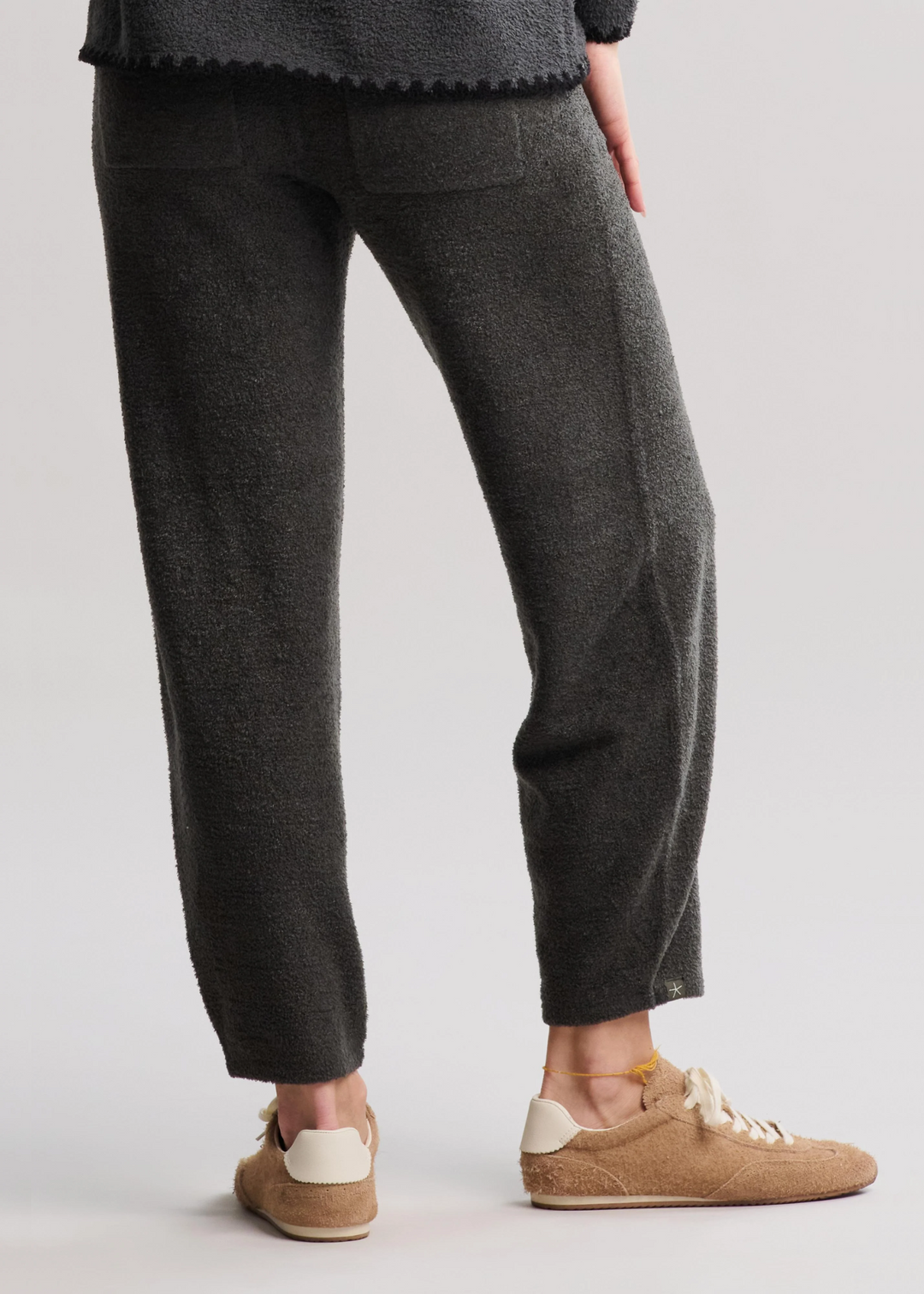 Woman turned around wearing dark green/grey barrel pants with tan sneakers in front of a white background