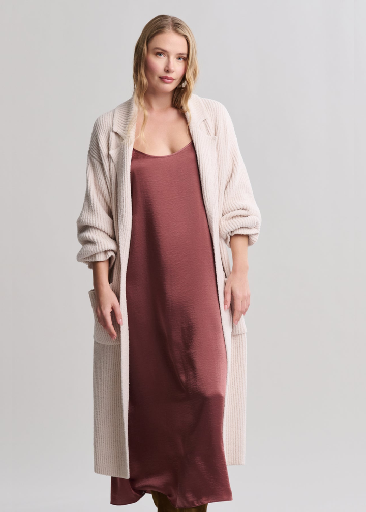 Woman wearing a long cream coat over a mauve maxi dress standing in front of a grey background