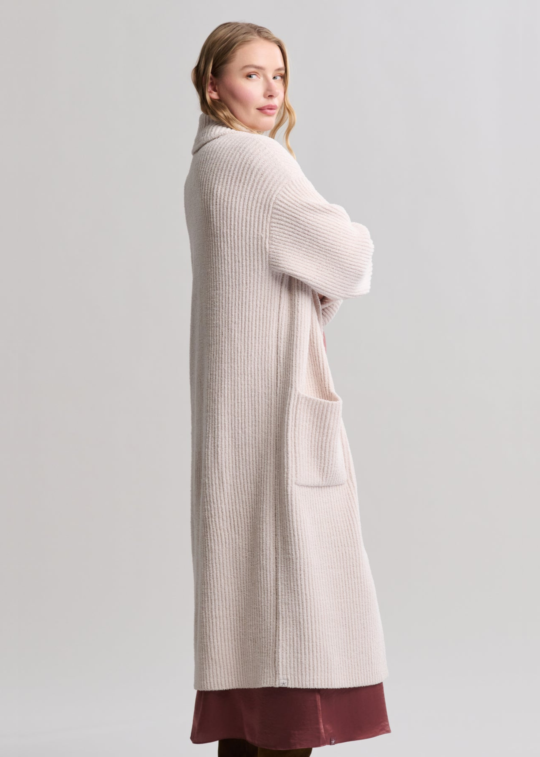 Woman turned to the side wearing a long cream coat over a mauve maxi dress standing in front of a grey background