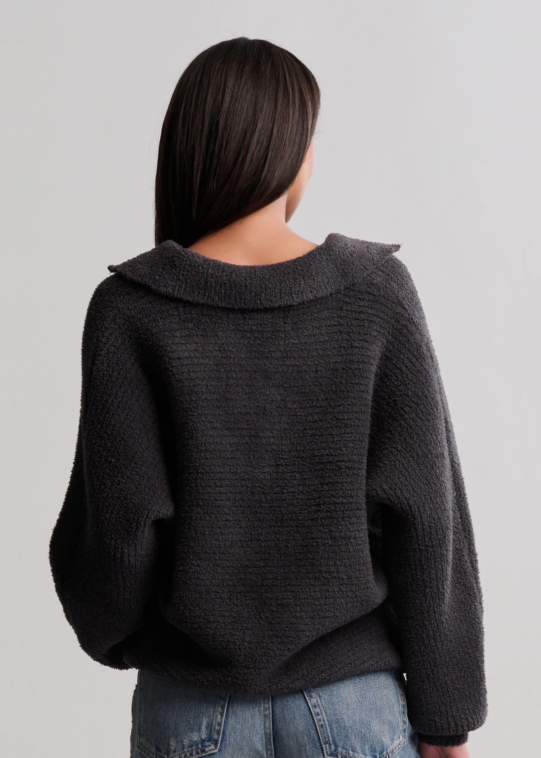 Woman turned around wearing a black collared pullover and jeans standing in front of a white background