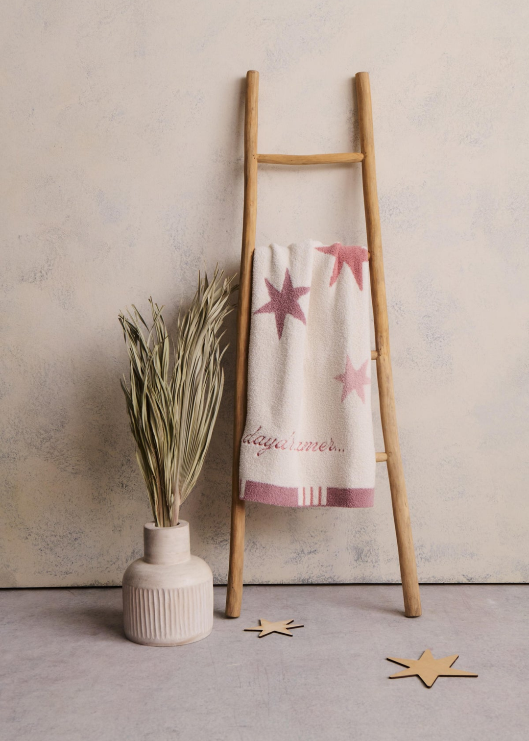 Pink blanket with stars and a striped border and "daydreamer..." pink embroidery hanging on a wooden ladder next to a plant
