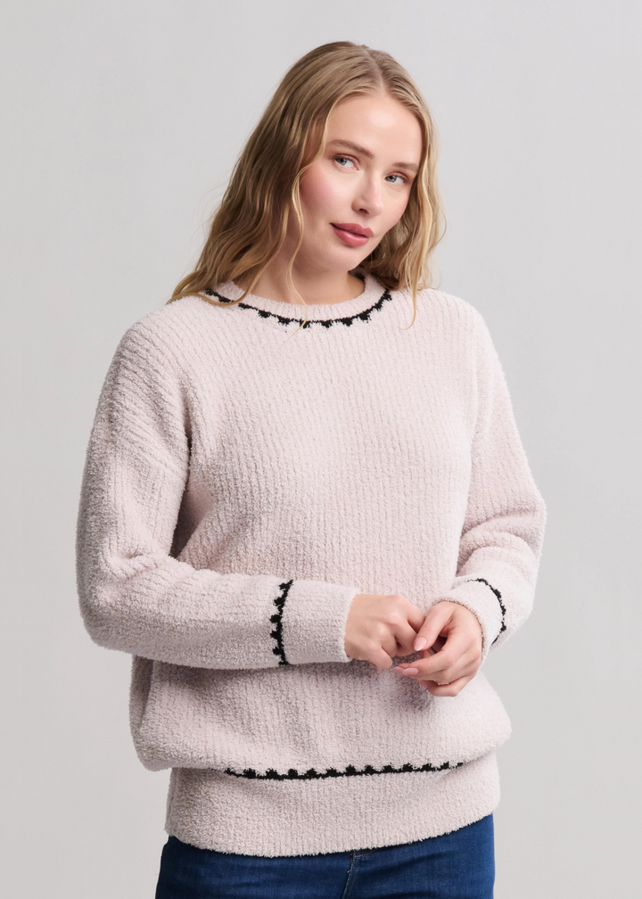 Woman wearing a cream colored sweater with black stitching and jeans standing in front of a white background