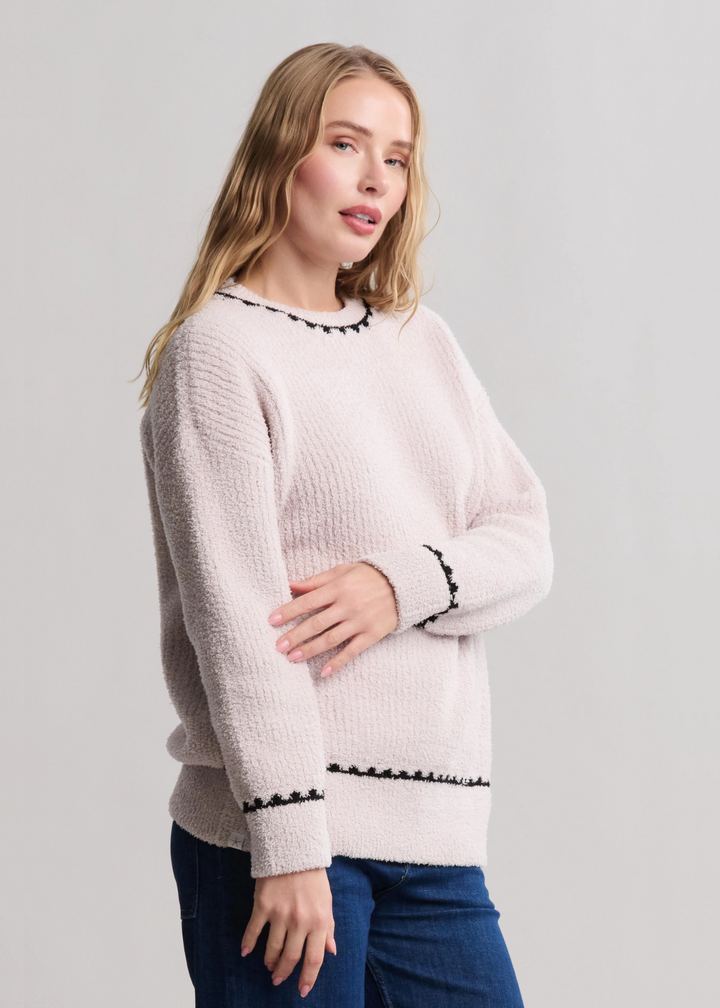 Woman turned to the side wearing a cream colored sweater with black stitching and jeans standing in front of a white background