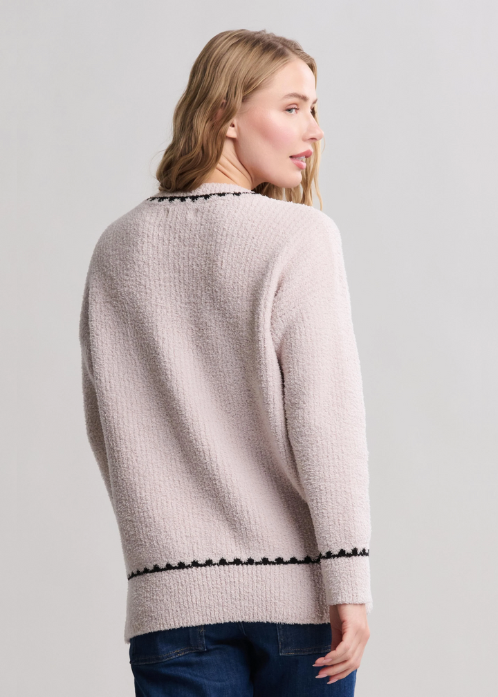 Woman turned around wearing a cream colored sweater with black stitching and jean standing in front of a white background