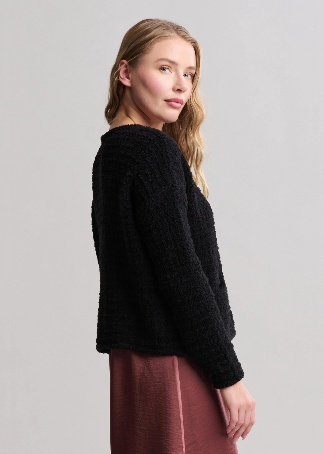 Woman turned to the side wearing a textured button front black cardigan with a mauve skirt standing in front of a white background