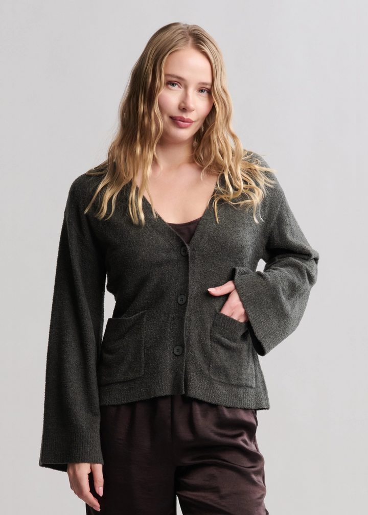 Woman wearing a dark green button front cardigan with brown pants standing in front of a white background