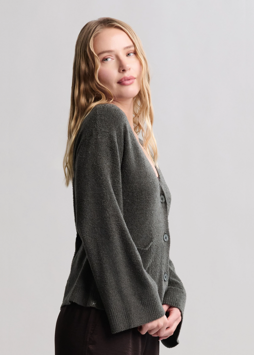 Woman turned to the side wearing a dark green button front cardigan with brown pants standing in front of a white background