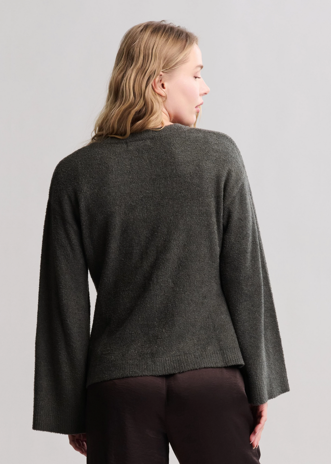 Woman turned around wearing a dark green button front cardigan with brown pants standing in front of a white background