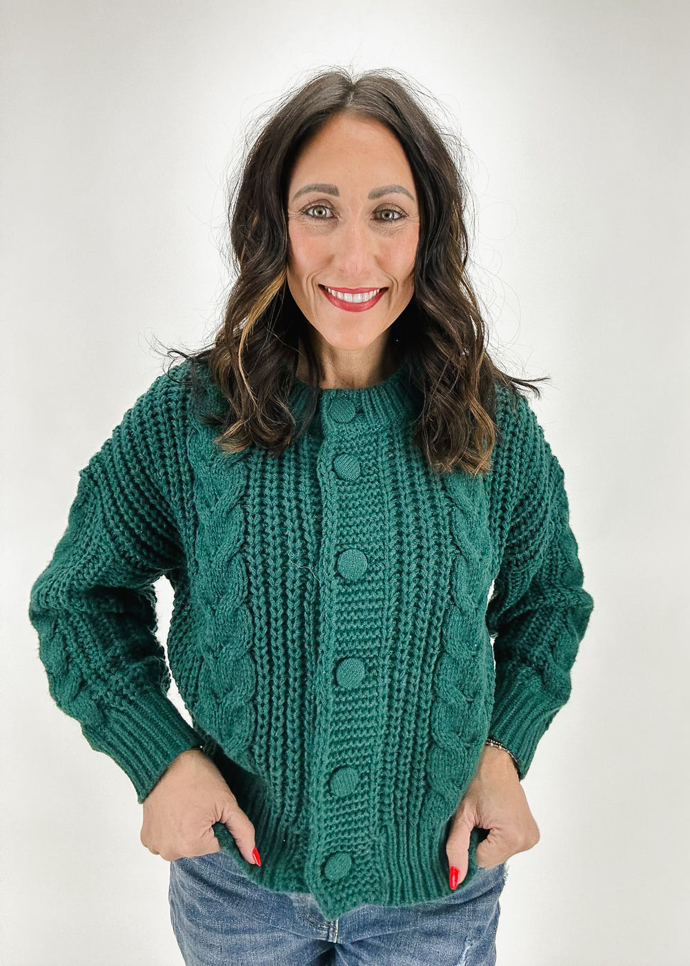 A woman wearing a chunky green cable-knit sweater with button detailing and long sleeves, styled with jeans for a cozy fall or winter look.