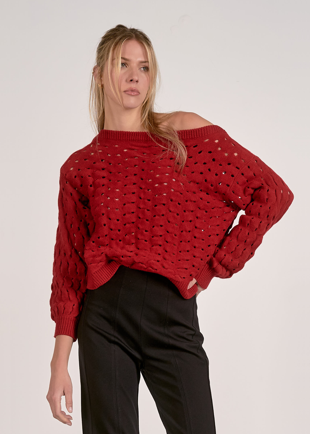 Woman wearing a red off the shoulder open knit sweater with black pants against a grey background