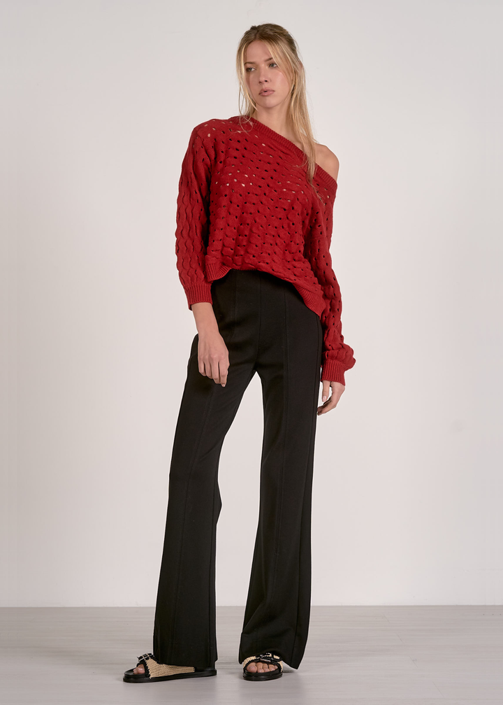 Woman wearing a red off the shoulder open knit sweater with black pants against a grey background