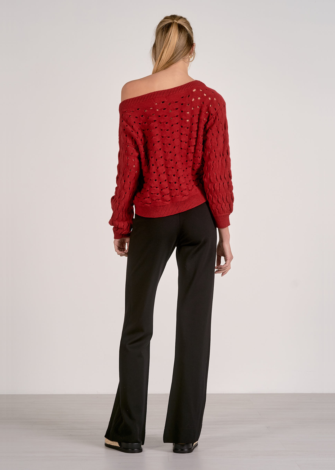 Woman turned around wearing a red off the shoulder open knit sweater with black pants against a grey background