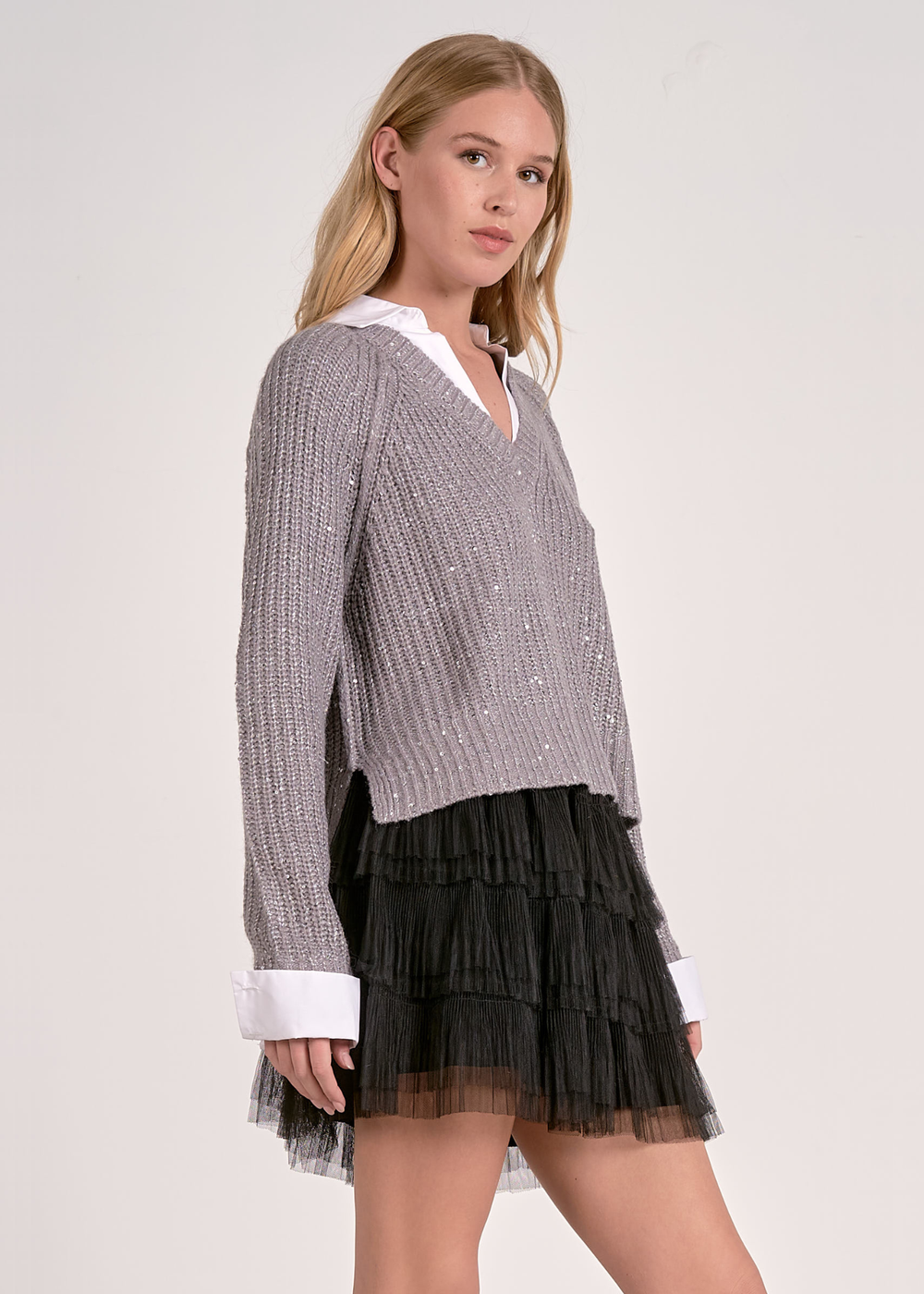 Woman turned to the side wearing a grey sparkly sweater layered over a white button down with a black mesh skirt