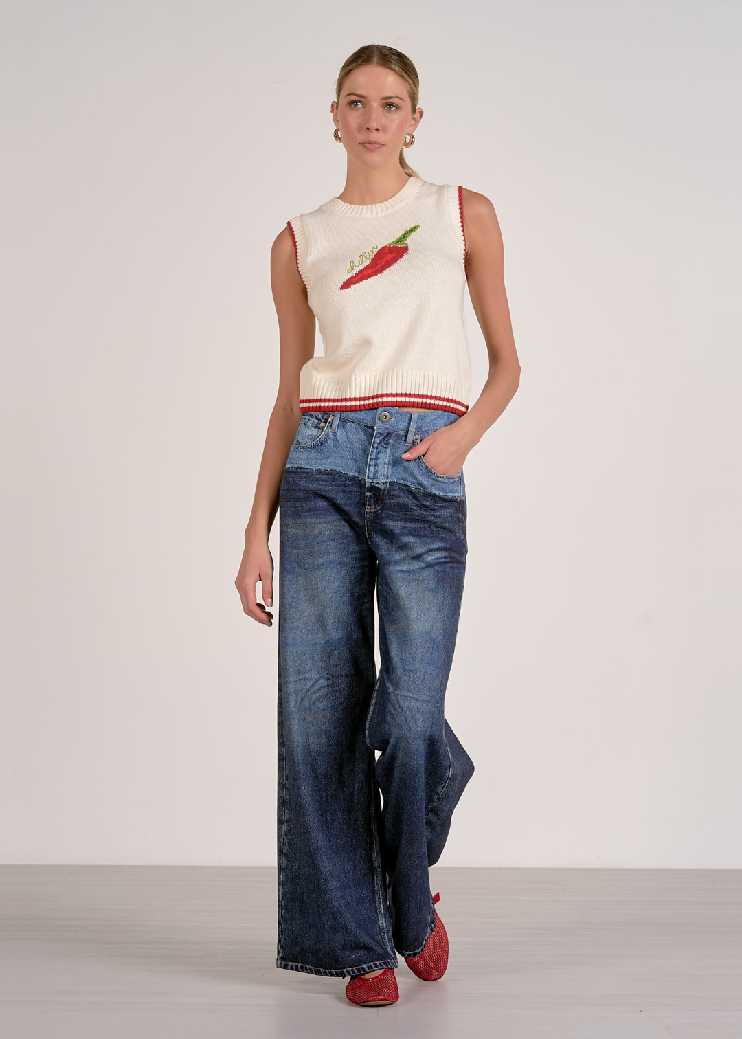 Woman with hand in pocket wearing a white sleeveless knit top with a red pepper and "chillin" graphic and jeans