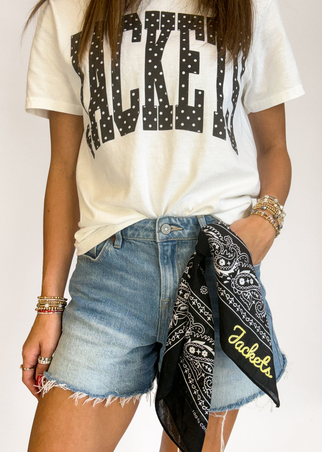 A person wearing a white graphic T-shirt with large black polka-dot block letters spelling 'JACKETS', light-wash denim shorts, and a black bandana with gold script and paisley details tied around the waist. The background is a plain light gray studio backdrop.