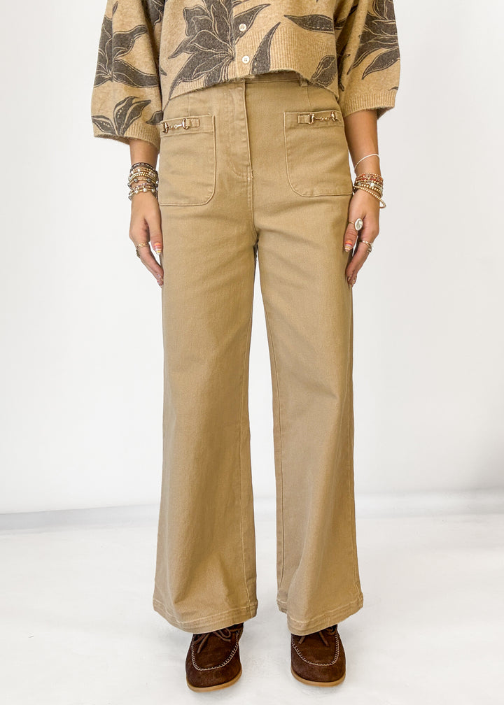 A person wearing tan wide-leg pants with front patch pockets, paired with a patterned beige and brown top and brown shoes, standing against a plain light background.
