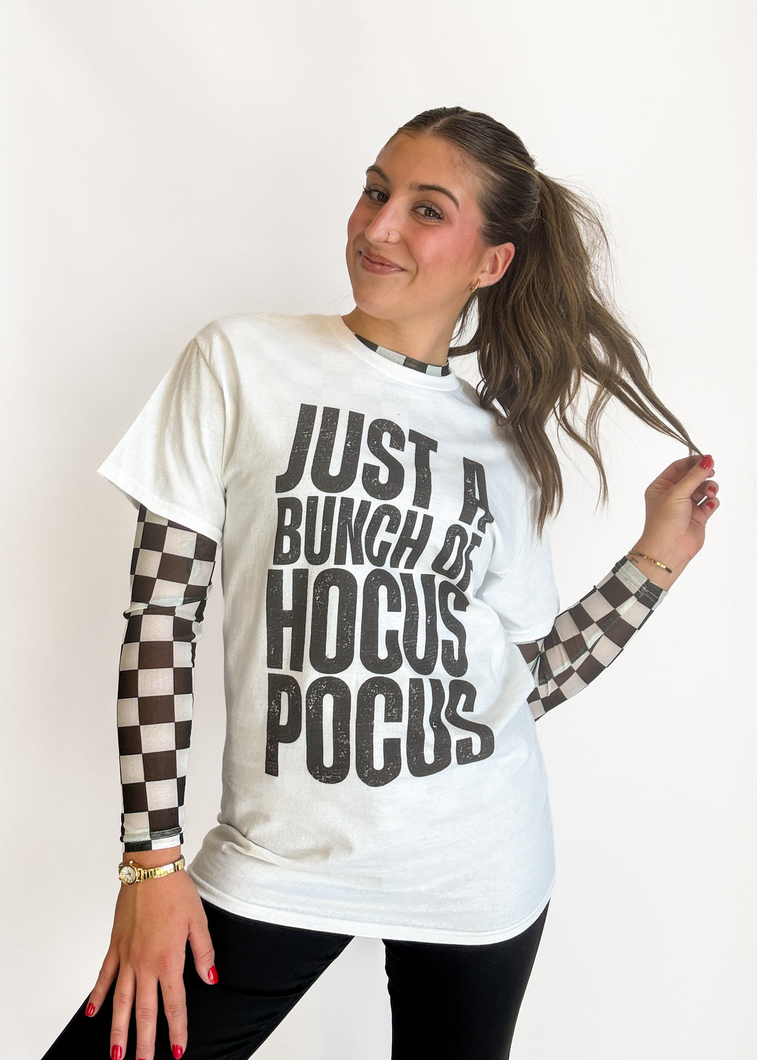 A woman modeling a white graphic t-shirt with black text reading 'JUST A BUNCH OF HOCUS POCUS' layered over black-and-white checkered long sleeves, against a plain white background.