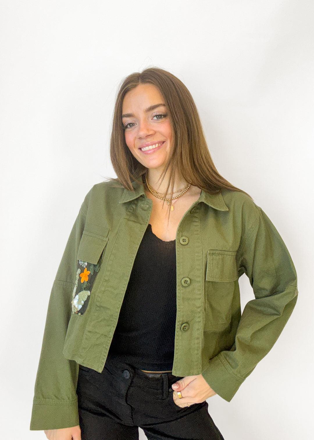 A woman wearing an olive green military-style utility jacket with embroidered patches on the chest, layered over a black top and black pants, standing against a plain white background.