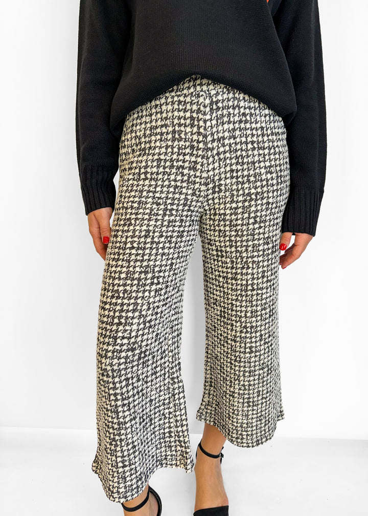 A person wearing black and beige houndstooth wide-leg cropped pants with a black sweater and black heels, shown from the chest down against a plain white background.