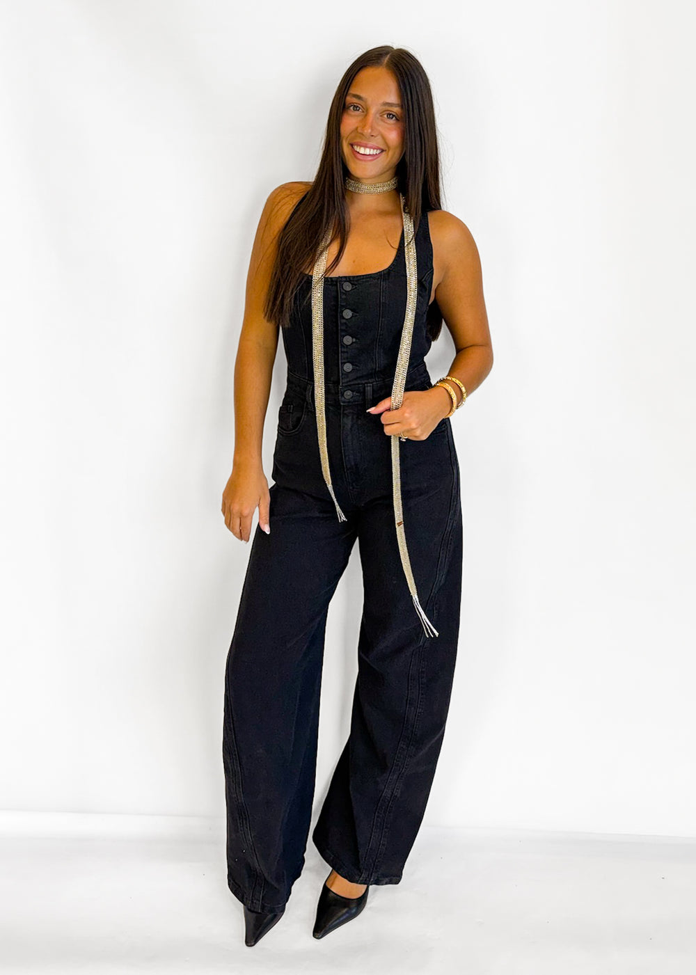 A woman modeling a black denim jumpsuit with wide legs and front buttons, styled with a beige measuring tape draped over her shoulders, set against a plain white studio background.