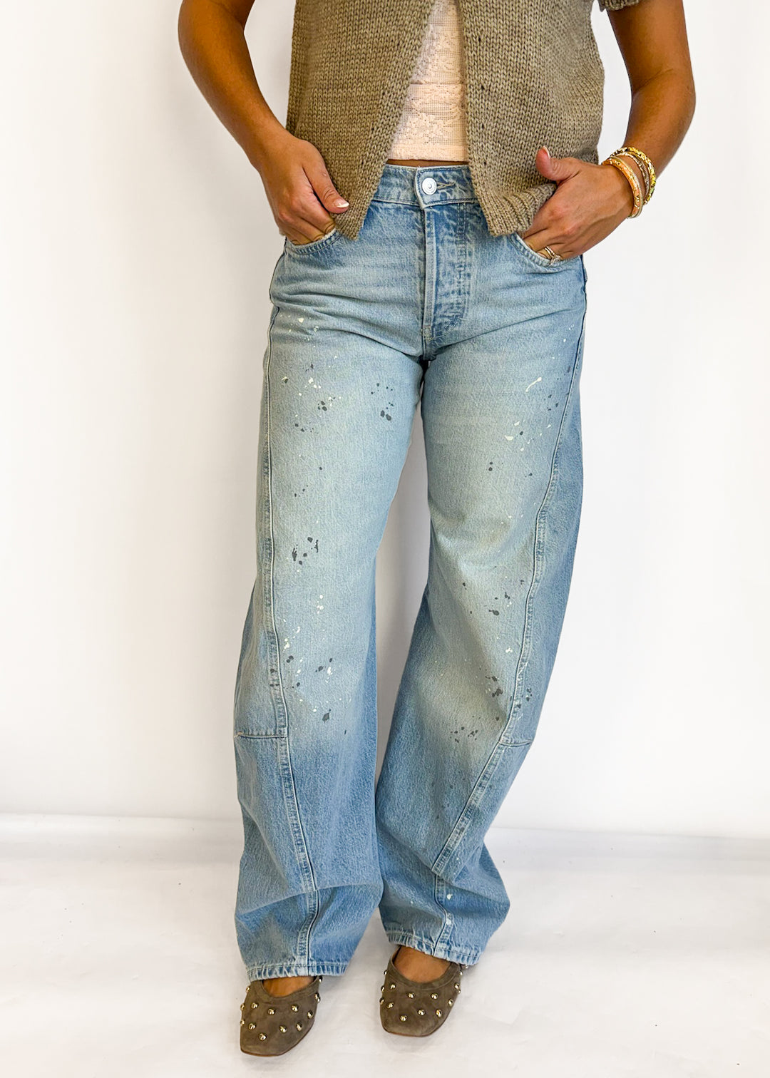 A person wearing light-wash blue denim jeans with small black speckled details, styled with a beige knit vest, a cream top, and brown studded shoes against a plain white studio background.