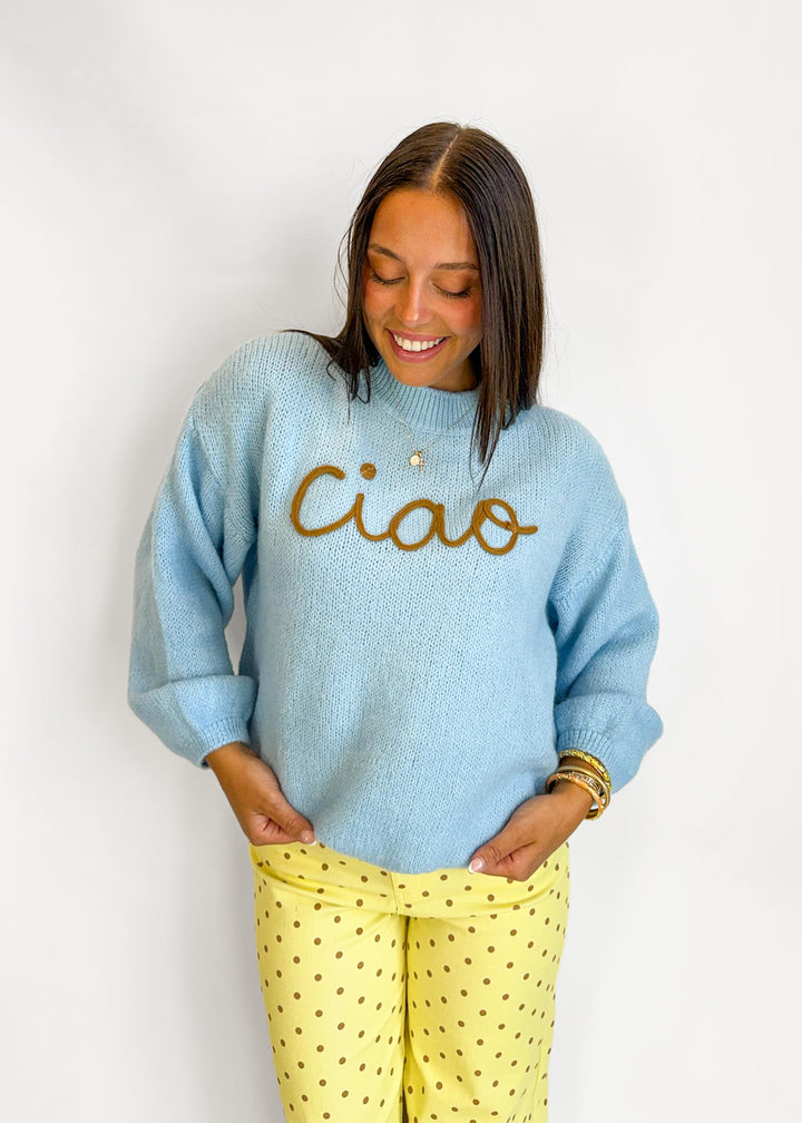 A woman wearing a light blue fuzzy knit sweater with 'Ciao' embroidered in brown across the chest, paired with yellow pants with small brown polka dots, standing against a plain white wall.