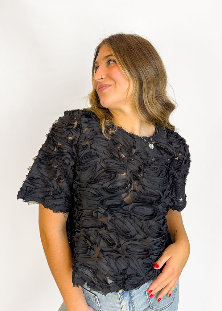 A woman wearing a black short-sleeve top with a heavily textured, ruffled, and shredded fabric design, paired with light blue jeans, photographed against a plain white background.