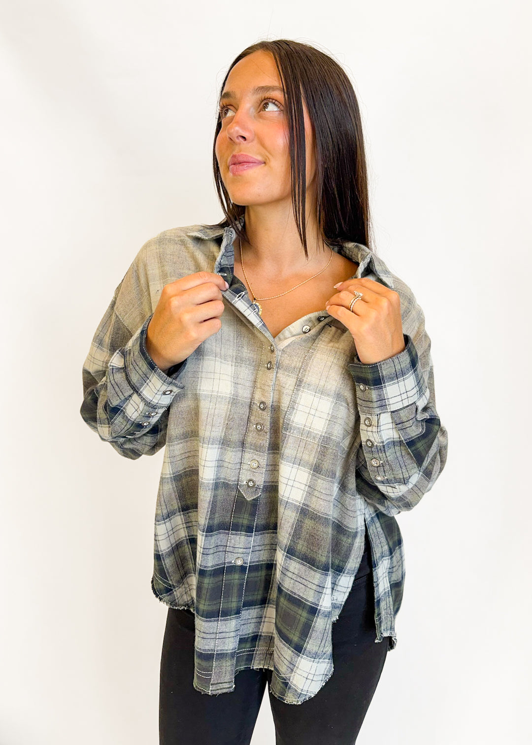 A woman wearing a long-sleeve plaid button-up shirt in muted gray, beige, and white tones, paired with black pants, posing against a plain white studio background.