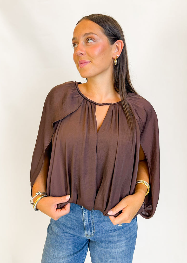 A woman wearing a brown long-sleeve blouse with a keyhole neckline and blue jeans, standing against a plain white background.