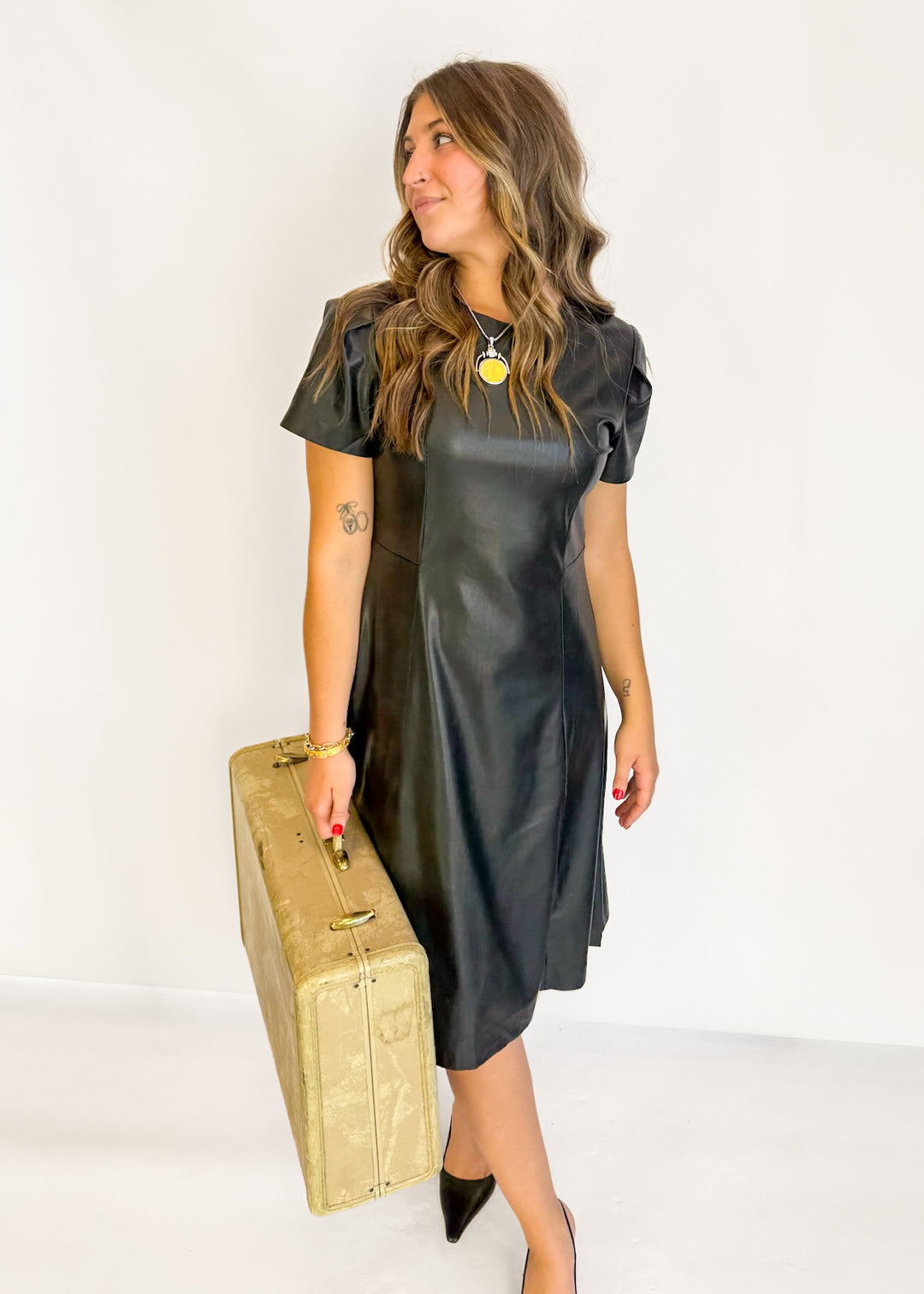 A woman wearing a black faux-leather short-sleeve dress and holding a vintage gold suitcase, standing against a plain white studio background.