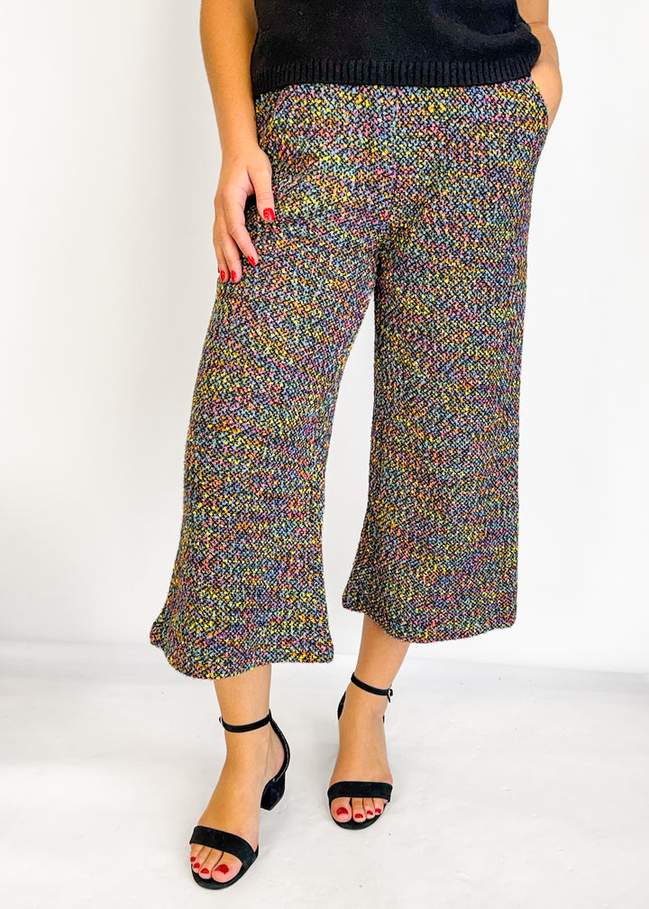 A model wearing a pair of cropped, wide-leg pants with a vibrant multicolor knit pattern, paired with black heeled sandals against a plain white studio background.