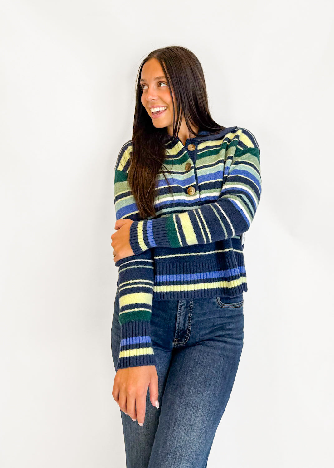 A woman wearing a striped knit cardigan sweater with blue, green, yellow, and white horizontal stripes, paired with dark blue jeans, standing against a plain white wall.