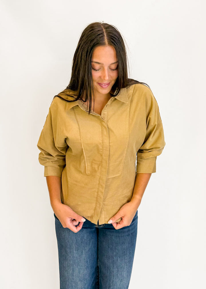 A woman wearing a tan long-sleeve button-up shirt with drawstrings and blue jeans, standing against a plain white background.