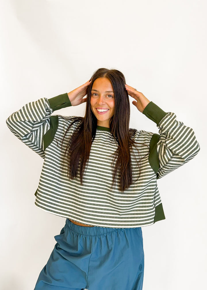 A woman posing in a cropped long-sleeve sweatshirt with green and white horizontal stripes and solid green ribbed cuffs and shoulders, paired with blue sweatpants against a plain white background.