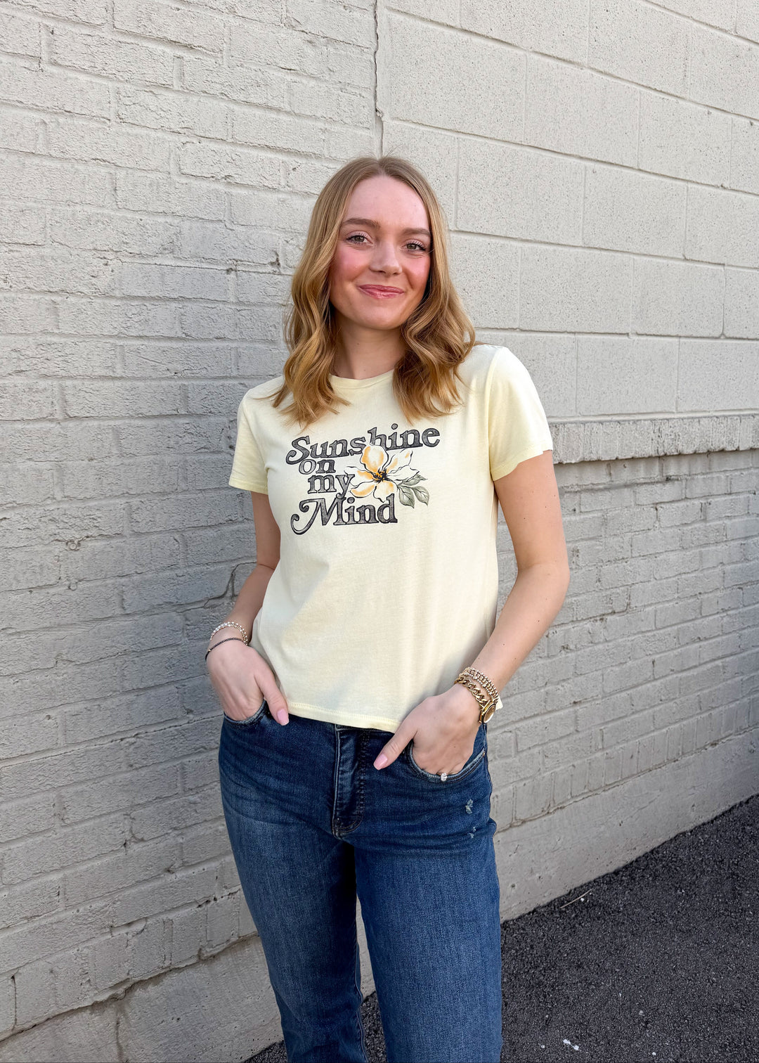 Person wearing a yellow t-shirt with text and graphics, standing against a light gray brick wall.
