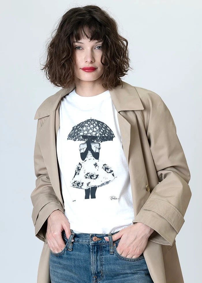Woman wearing a beige trench coat over a white t-shirt with a graphic design, standing against a plain background.