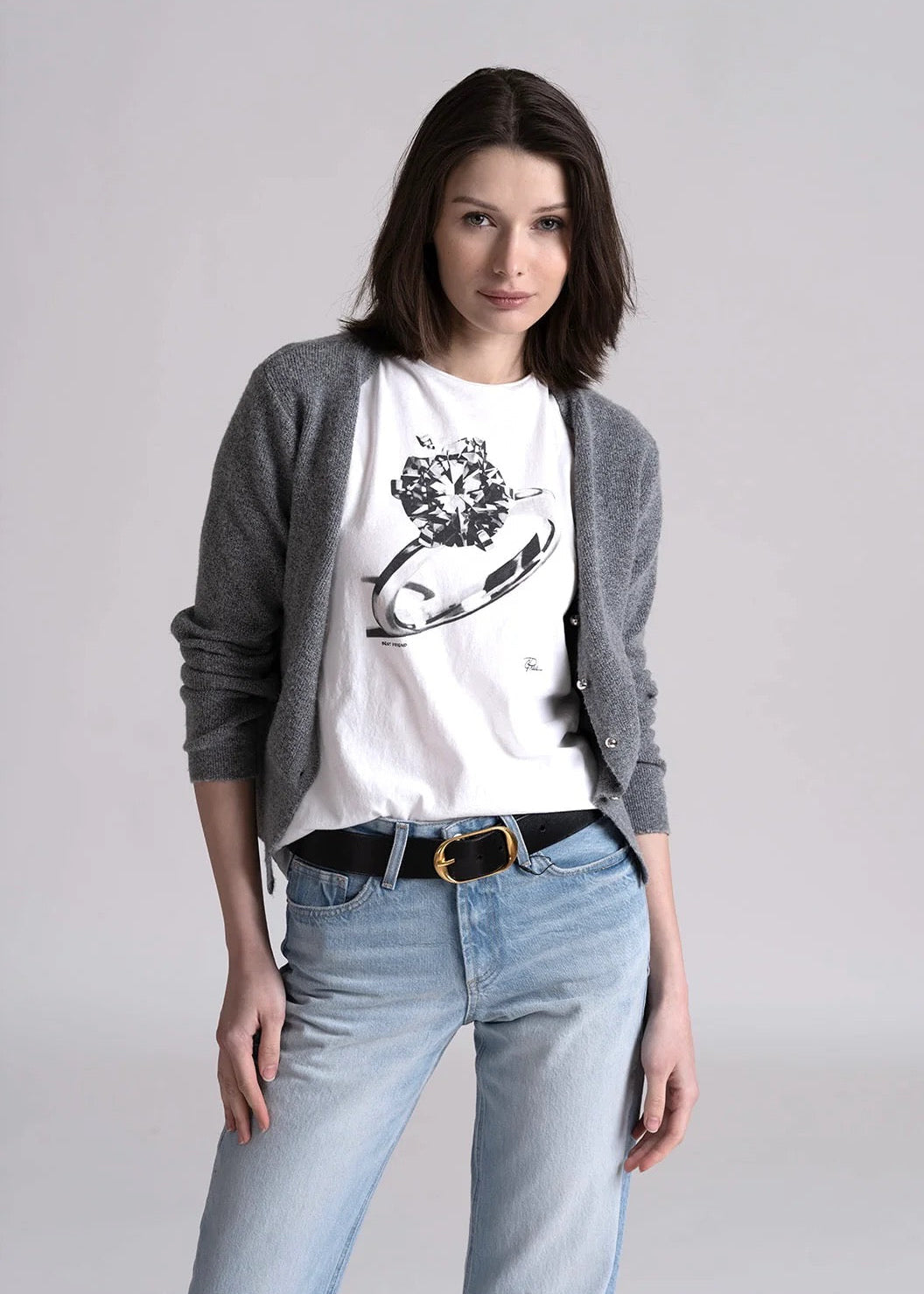 Woman wearing a white t-shirt with a graphic design, gray cardigan, and blue jeans on a plain background