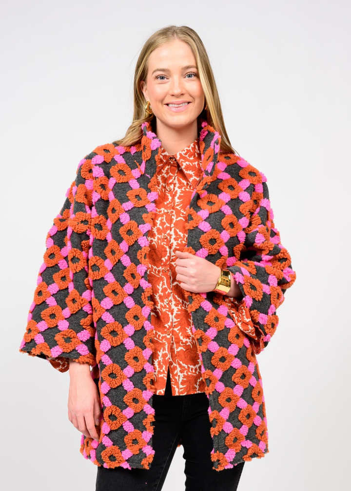 Woman wearing a pink, grey, and organe coat over an orange top and black pants in front of a white background
